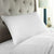 A crisp, white Downlite® DOWNLITE 50/50 White Goose Down & Feather Pillow | Featured in Many Hotels sits against a tufted, light gray headboard. The bed is made with matching white bedding. To the right of the headboard, there is a modern nightstand holding a dark blue ceramic lamp with a white shade and a small glass object. The background is bright and well-lit.