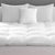 A monochromatic, minimalist styled bed with plush white bedding, featuring a Hollander Chaps Baffle Box Feather Bed, made in the USA, and an arrangement of decorative pillows with varying patterns in a serene grey-scale setting.