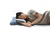 A person with shoulder-length dark hair is lying on their side, resting their head on an OPULENCE Glacier Ergonomic Memory Foam Pillow | Contour by Opulence. They are wearing a light beige shirt and appear to be asleep or resting against a plain white background, enjoying the neck support provided.